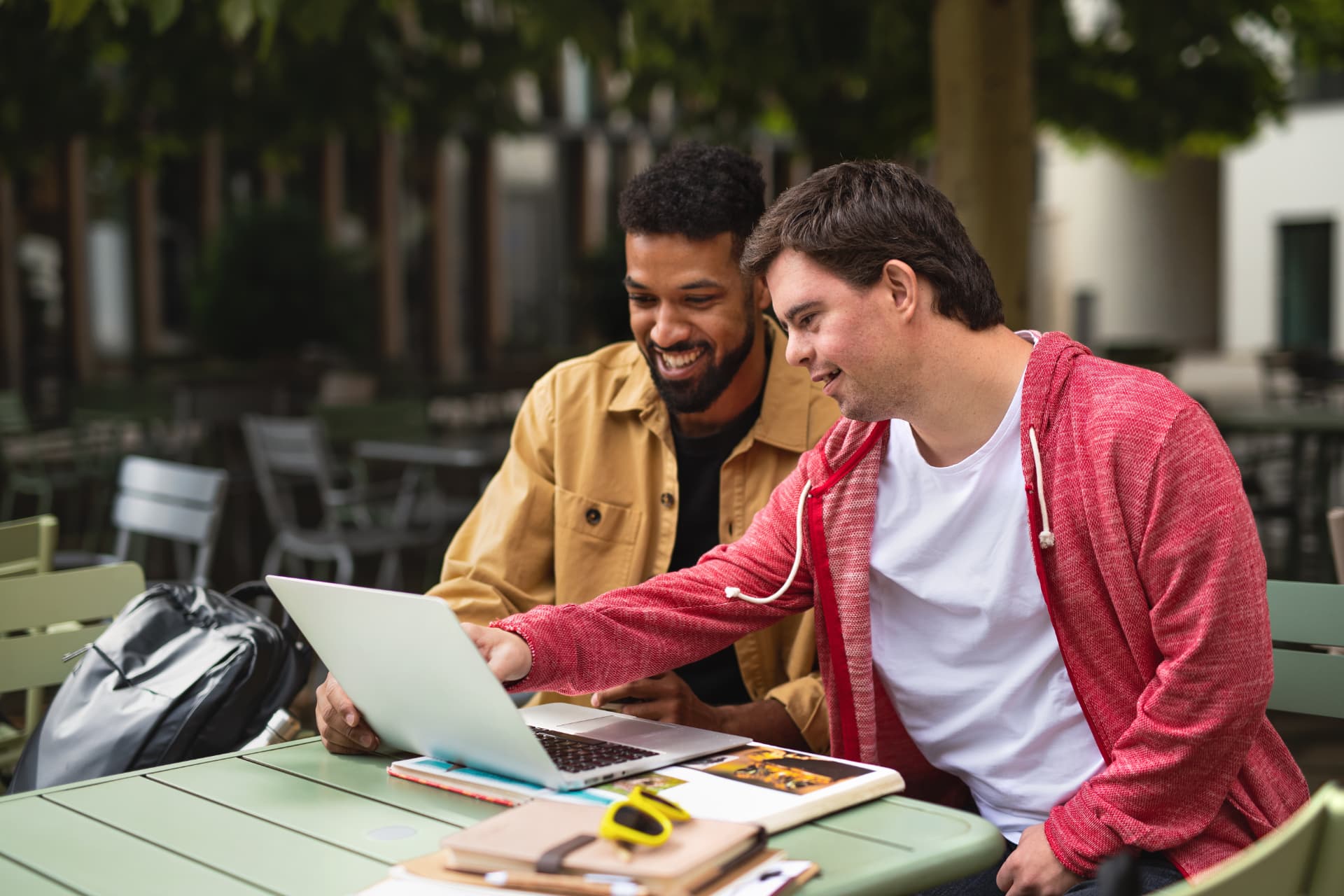 Two young men smiling while using a laptop together at an outdoor table