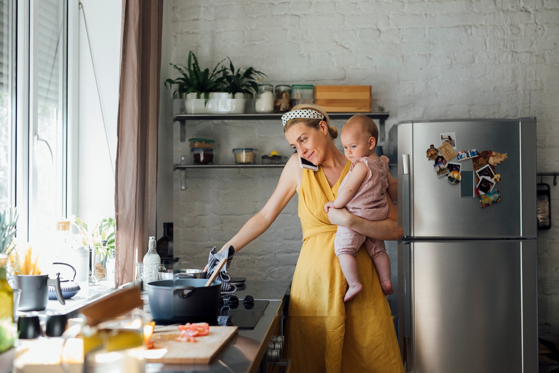 Mother holding baby while cooking and talking on the phone in kitchen