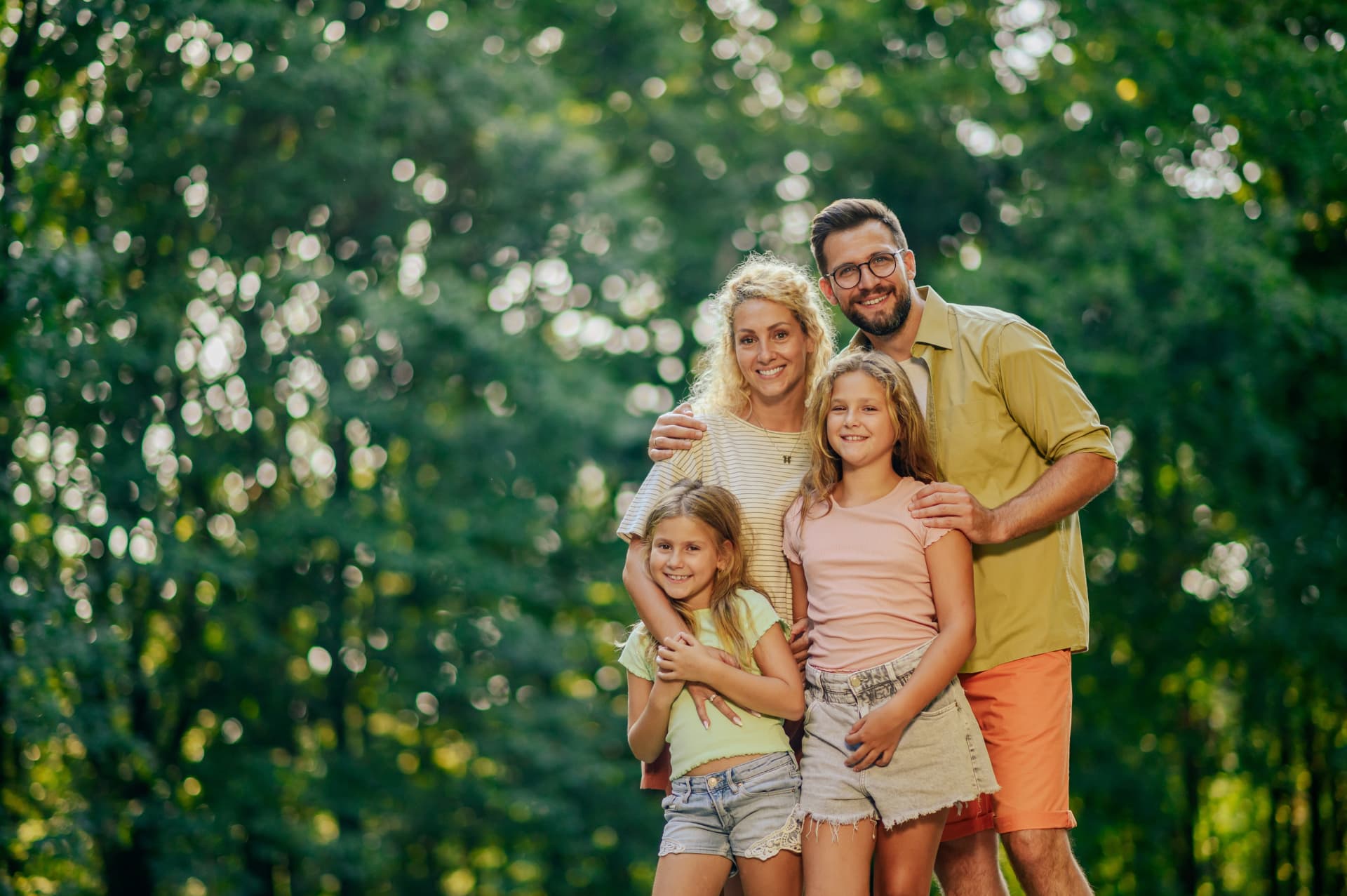 Smiling family of four with two young daughters standing together outdoors in a green park